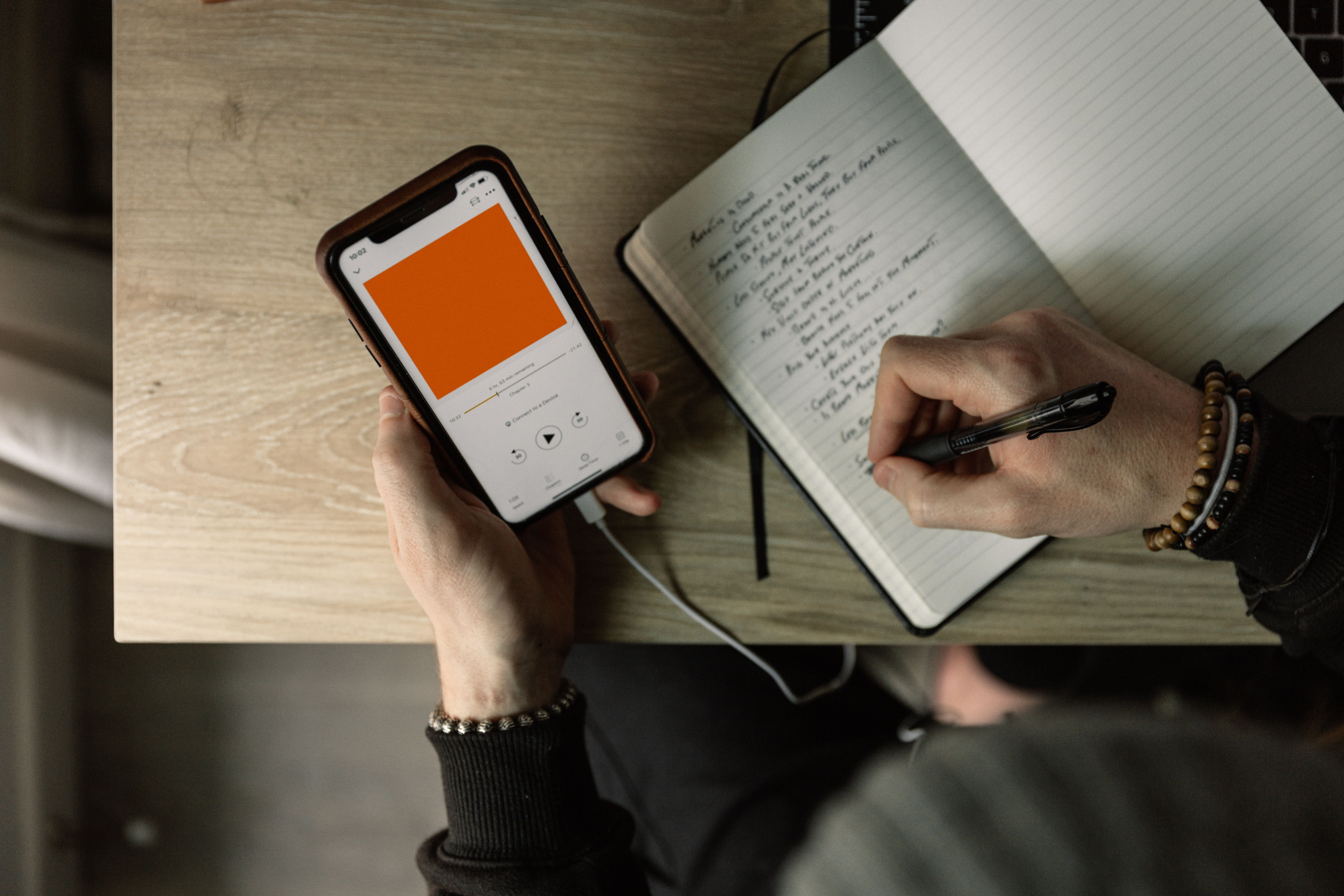 Uma pessoa segurando o celular na mesa enquanto escreve em um caderno